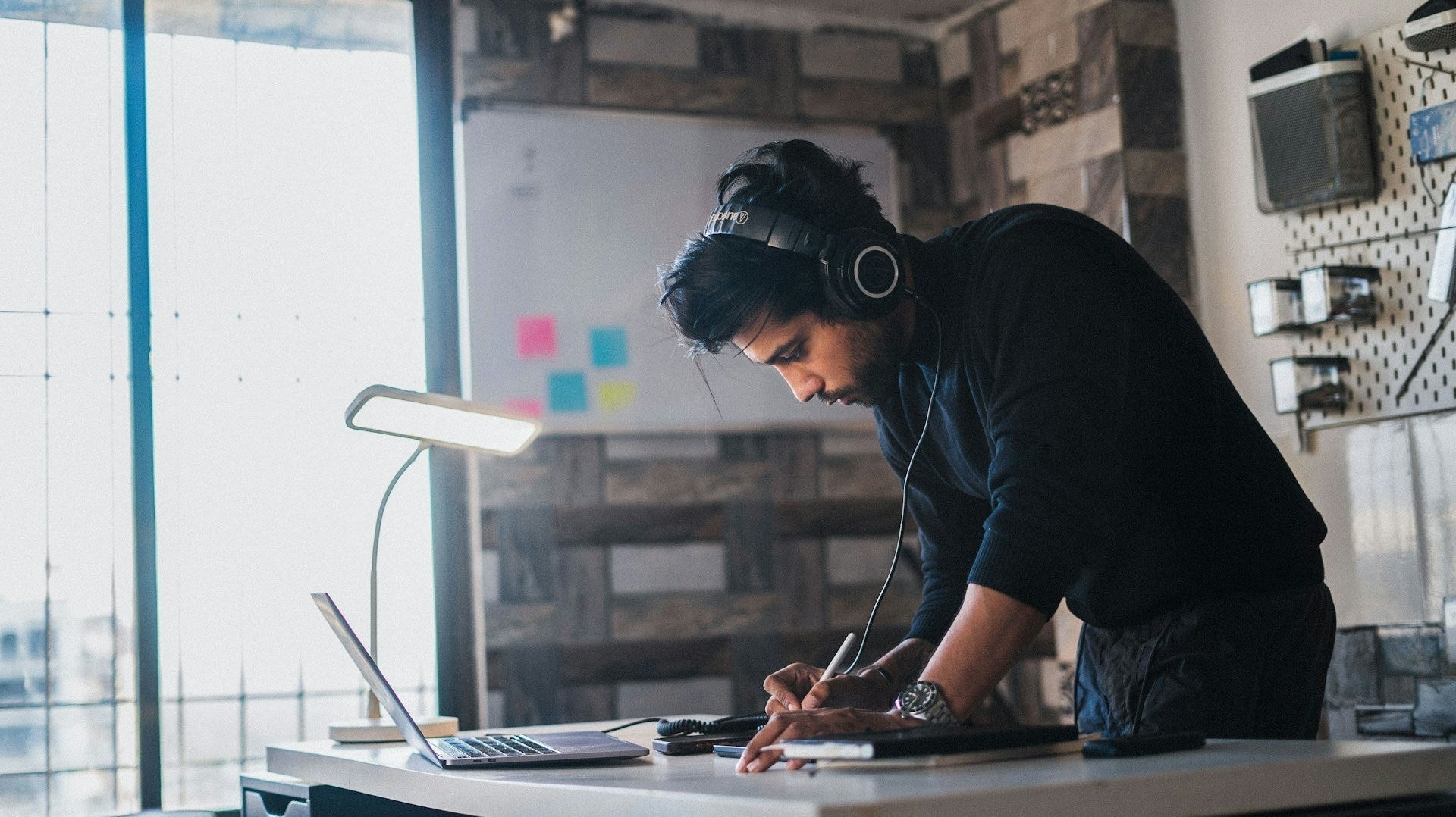 Man listening to music for work focus while writing at a desk with a laptop, headphones, and bright task lighting. Photo by Puneet Kaul via Unsplash