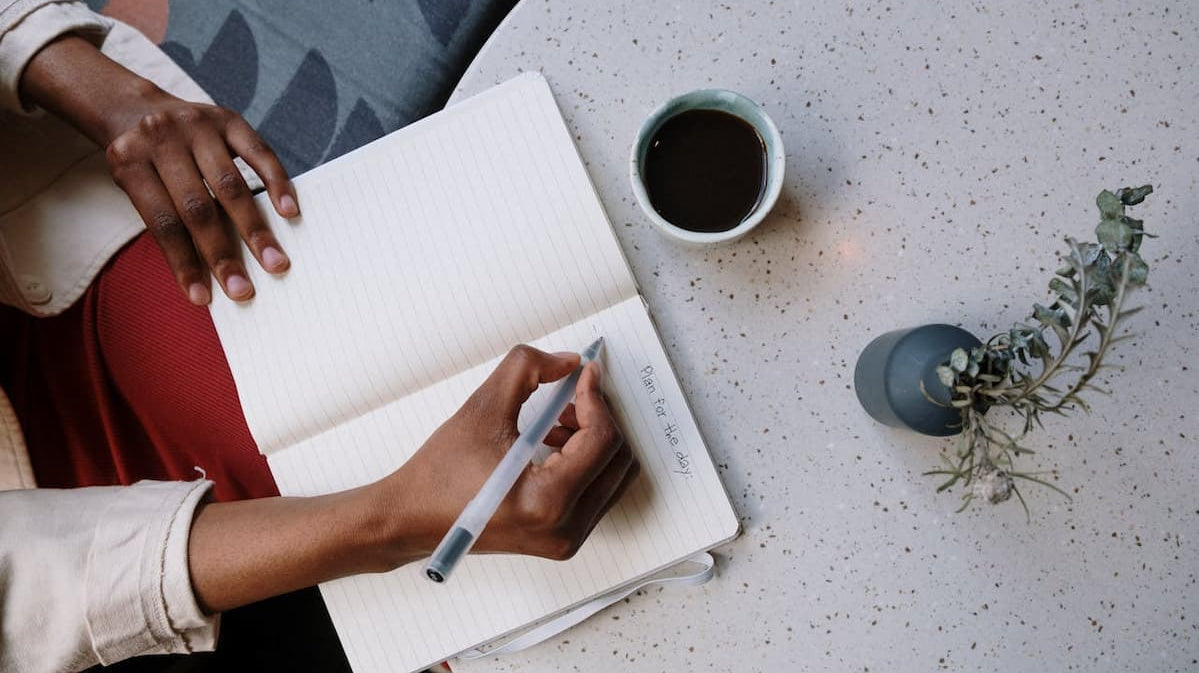 Person writing in a notebook with coffee and plant nearby, promoting a calm, productive work environment. Image by Cottonbro Studio via Pexels