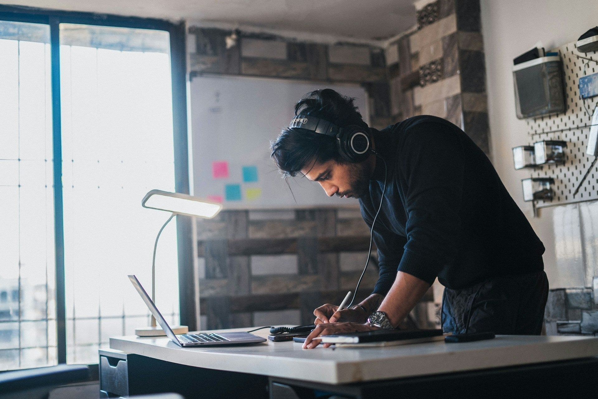 Man listening to music for work focus while writing at a desk with a laptop, headphones, and bright task lighting. Photo by Puneet Kaul via Unsplash