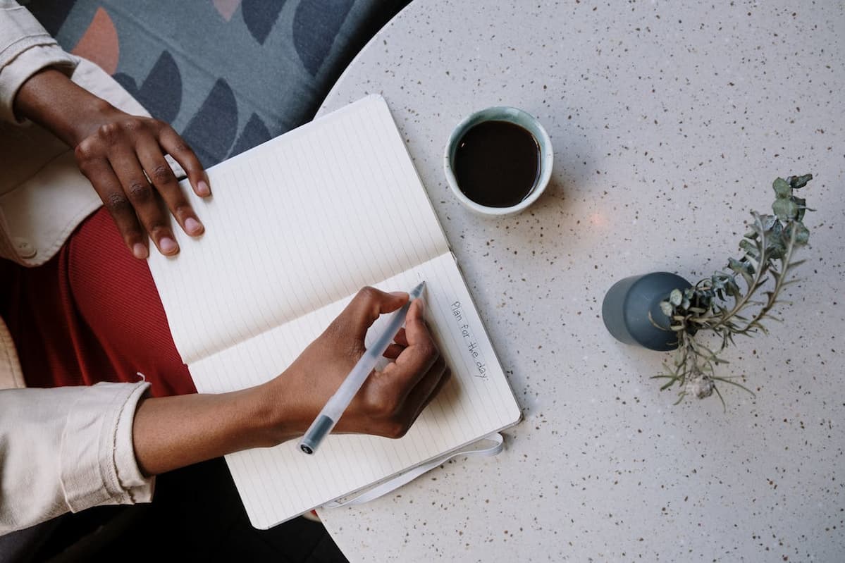 Person writing in a notebook with coffee and plant nearby, promoting a calm, productive work environment. Image by Cottonbro Studio via Pexels