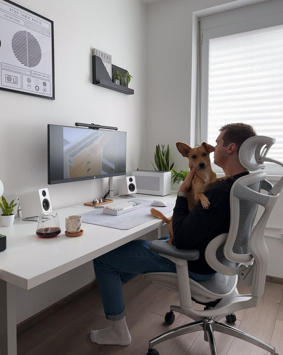 Man watching his computer while holding a dog and sitting on a white ergonomic chair.