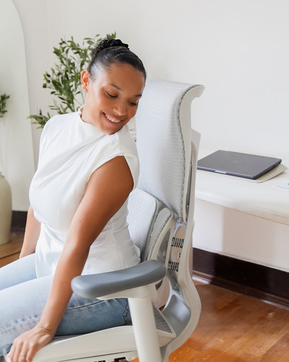 Woman sitting on a white ergonomic chair, smiling while looking at the back support feature.